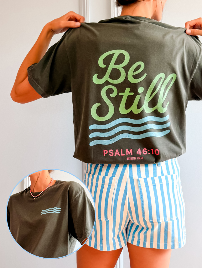 Be Still - Colorful Edition t-shirt in olive green with light blue text and waves, paired with striped shorts.