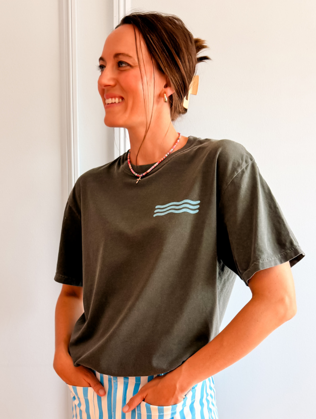 Be Still - Colorful Edition t-shirt in dark gray with blue wave design, worn by a smiling woman with hands in pockets.