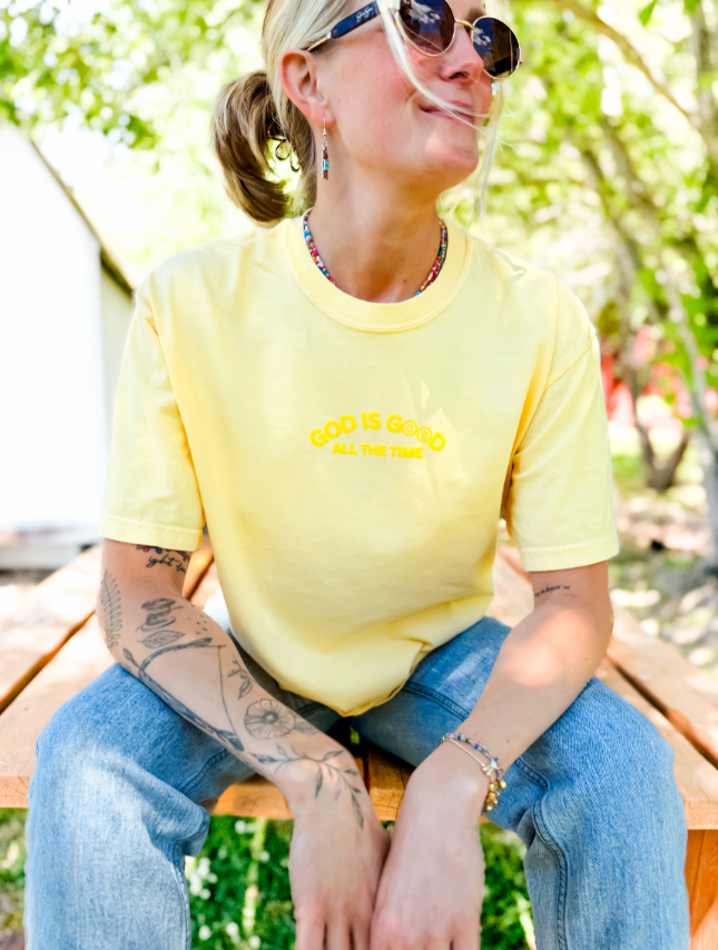 Yellow God Is Good Puff Tee worn by a person sitting on a wooden bench, surrounded by greenery.