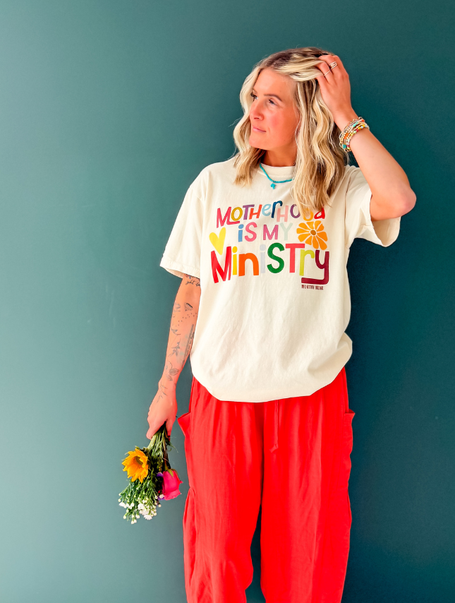 Motherhood is my ministry tee in cream, worn by a woman with flowers, paired with red pants against a green background.