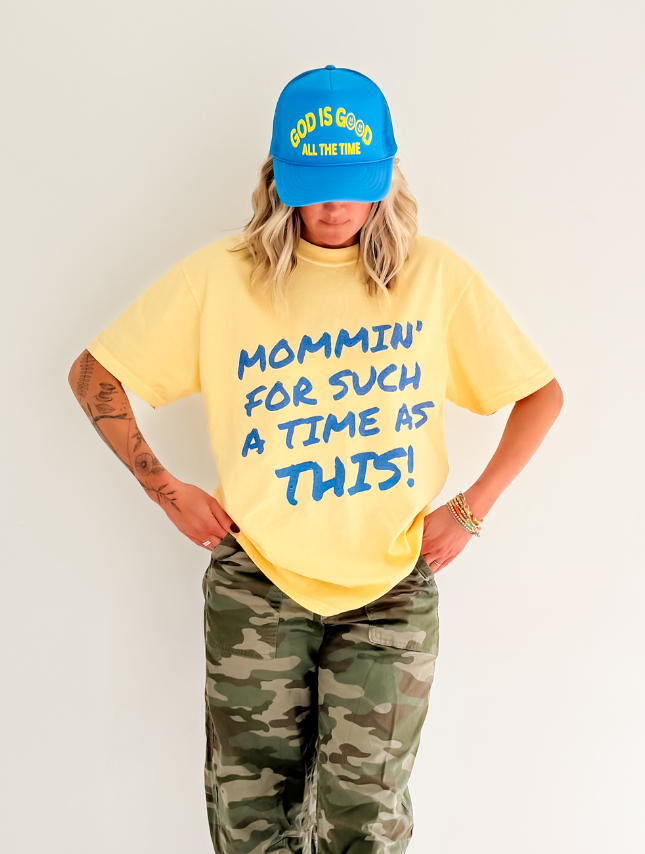 Yellow Mommin' Tee with blue text reading "Mommin' for such a time as this!" worn with camo pants and a blue cap.