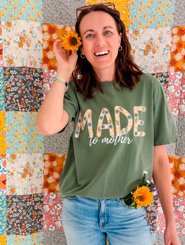 Green tee with "Made to Mother" text, worn by a woman holding a sunflower, against a colorful quilt background.