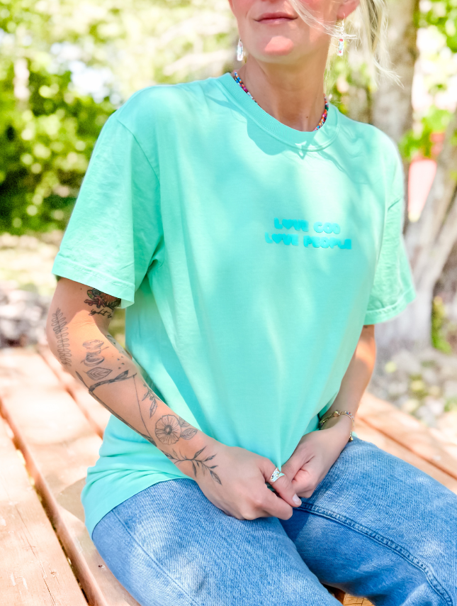 Mint green puff tee with "Love God Love People" text, worn by a person sitting outdoors on a wooden bench.