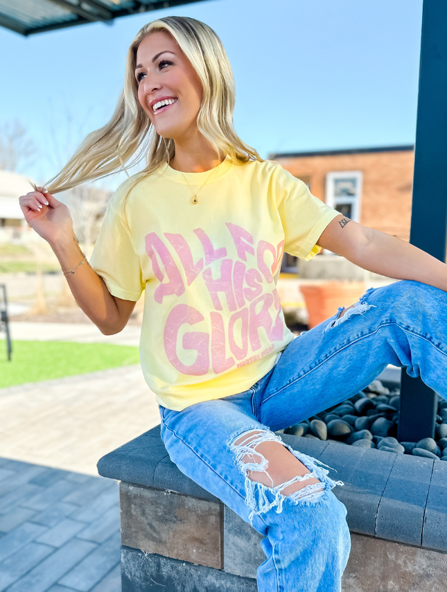 Yellow "All For His Glory" tee worn by a smiling woman, paired with ripped blue jeans, sitting outdoors.