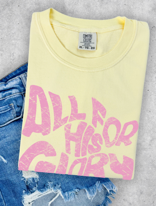 Yellow t-shirt with pink "All For His Glory" text, folded on a light gray surface next to denim shorts.