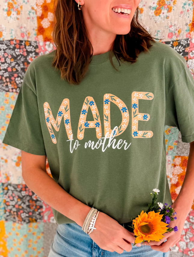 Green Made To Mother tee with floral design, worn by a woman holding a sunflower, against a colorful quilt background.