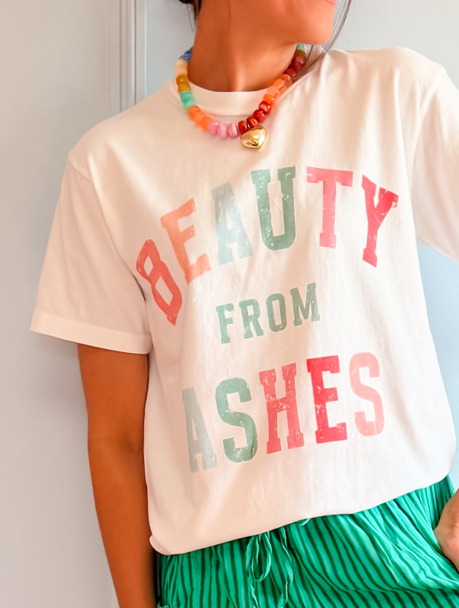 White tee with "Beauty From Ashes" in colorful letters, worn with green striped pants and a colorful beaded necklace.