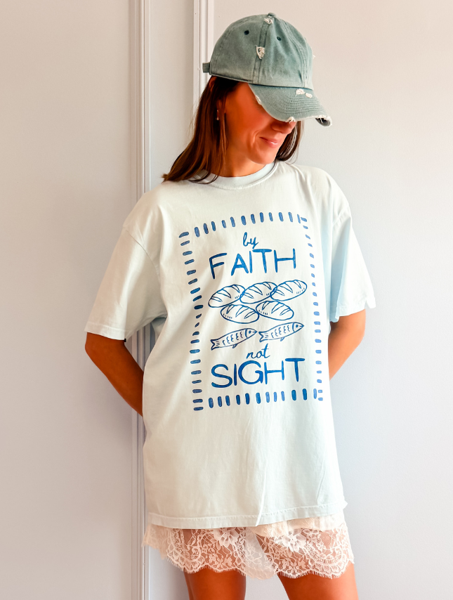 Light blue oversized tee with "By Faith Not Sight" graphic, worn with a green cap and white lace shorts.