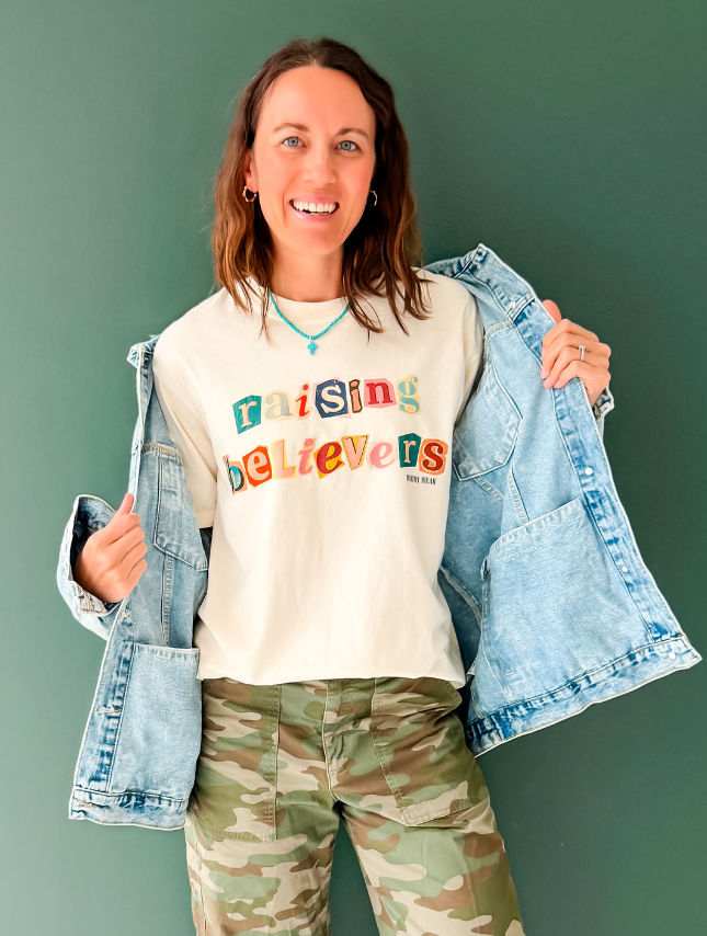 Raising Believers tee in cream, featuring colorful text, worn with a denim jacket and camo pants against a green background.