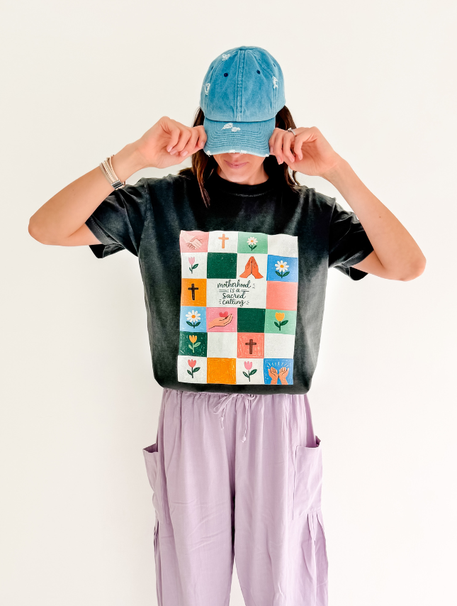 Sacred Calling Tee in black with colorful square graphic, worn by a model with a blue cap and lavender pants.