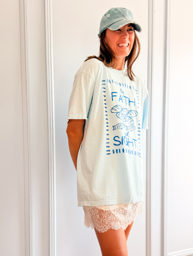 Light blue oversized tee featuring the phrase "By Faith Not Sight" with a graphic design, worn with a white lace skirt.