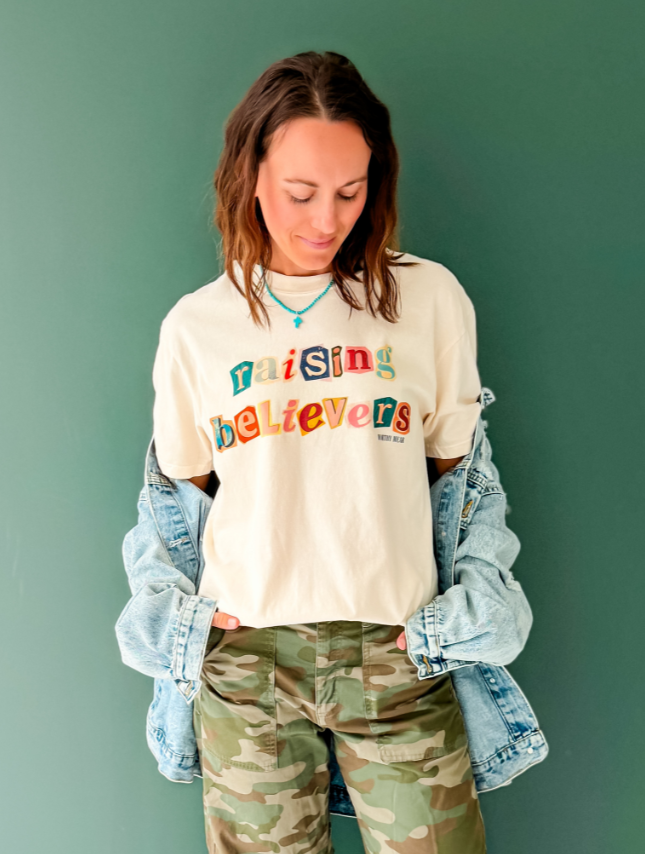 Raising believers tee in cream, featuring colorful text, worn with a denim jacket and camo pants against a green background.