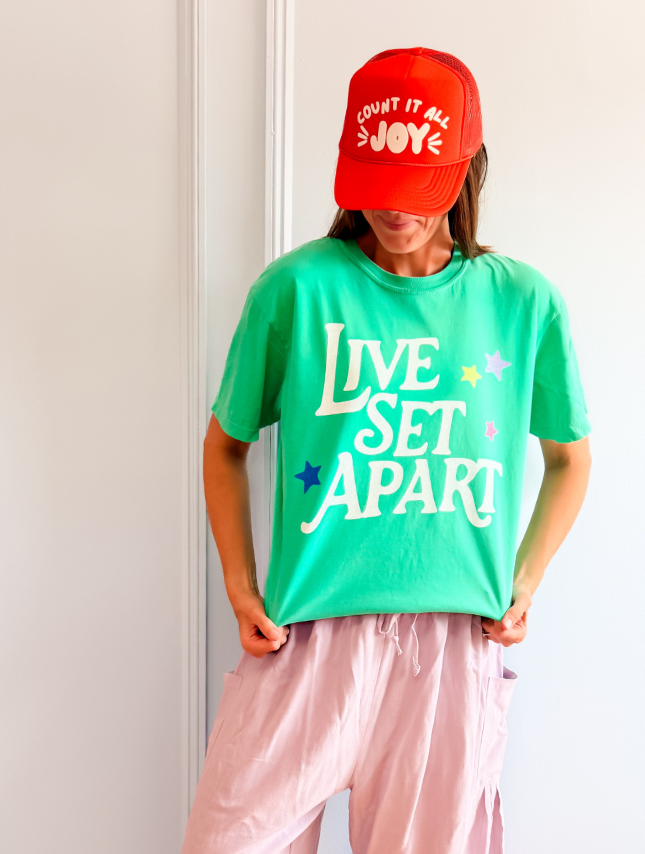Green "Live Set Apart" tee with colorful stars, worn by a person with a red cap and light pink pants.