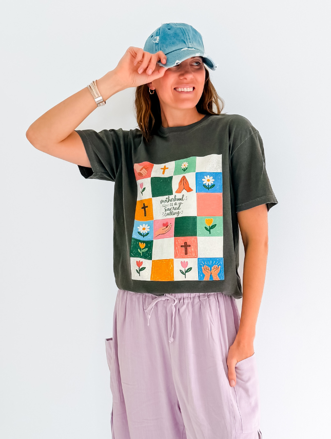 Sacred Calling Tee in dark gray with colorful graphic design featuring flowers and crosses, worn by a model with a blue cap.
