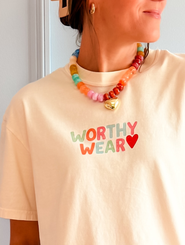 Cream-colored t-shirt featuring the text "Worthy Wear" in colorful letters, worn with a multi-colored bead necklace.