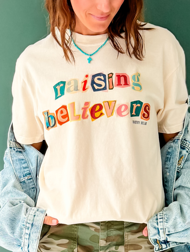 Raising believers tee in cream with colorful letters, worn with a denim jacket and layered necklace.