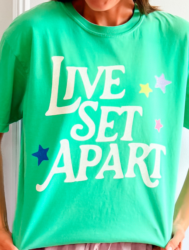 Green t-shirt featuring the text "Live Set Apart" in white, with colorful star accents, worn by a model.