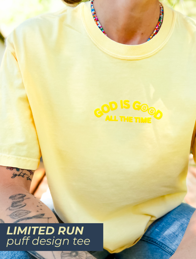 Yellow puff tee with "God Is Good All The Time" text, worn by a person with tattoos and colorful beads.